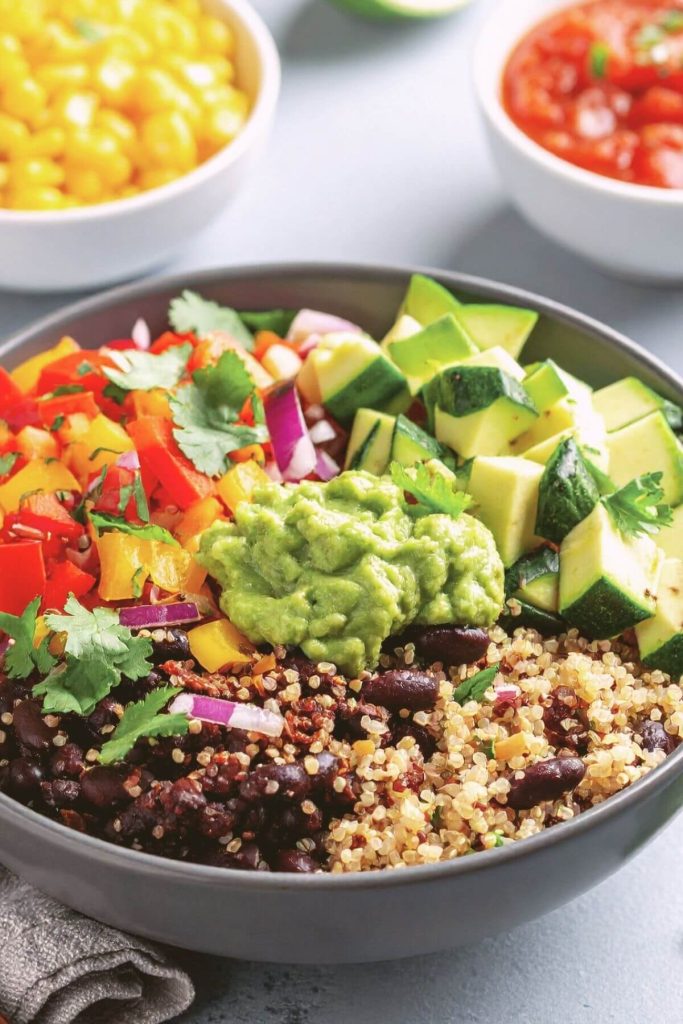 Vegan Burrito Bowl with Black Beans, quinoa, colorful peppers, zucchini, and guacamole with corn and salsa in small bowls