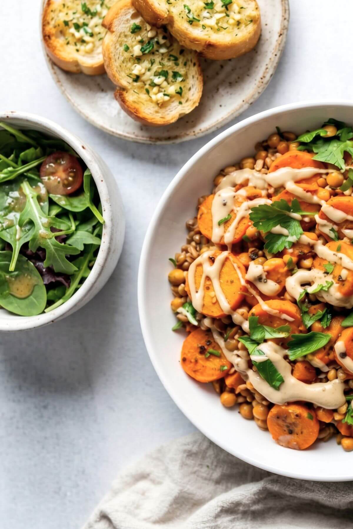 Roasted carrot and lentil salad with creamy tahini dressing being drizzled over top served wtih salad and garlic bread for meal